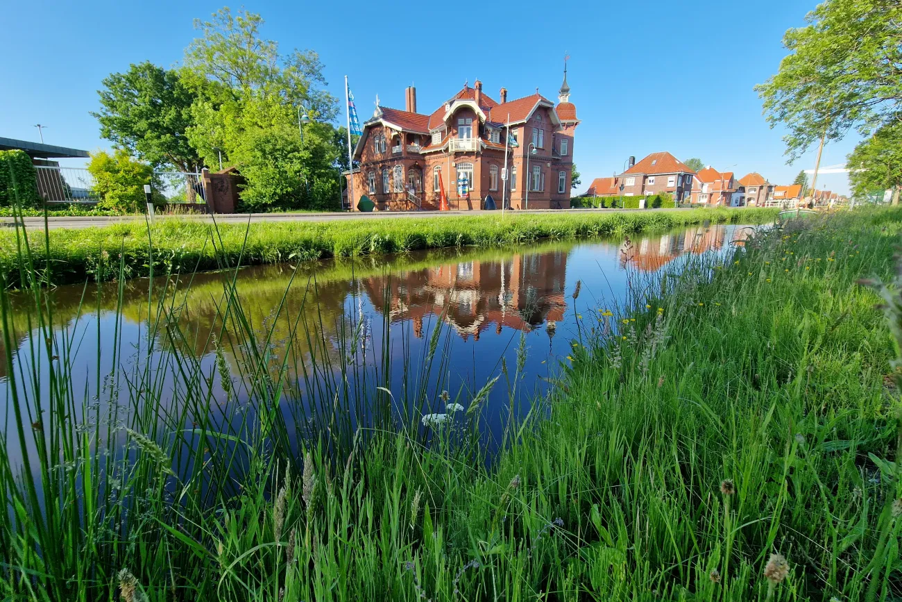 Rotes Backsteinmuseum spiegelt sich im Wasser des Rajenkanals hinter grünem Ufergras.