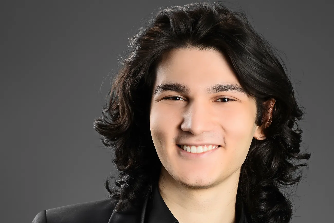 Lächelndes Porträt des Pianisten Kaan Baysal mit dunklem Haar vor grauem Hintergrund.
