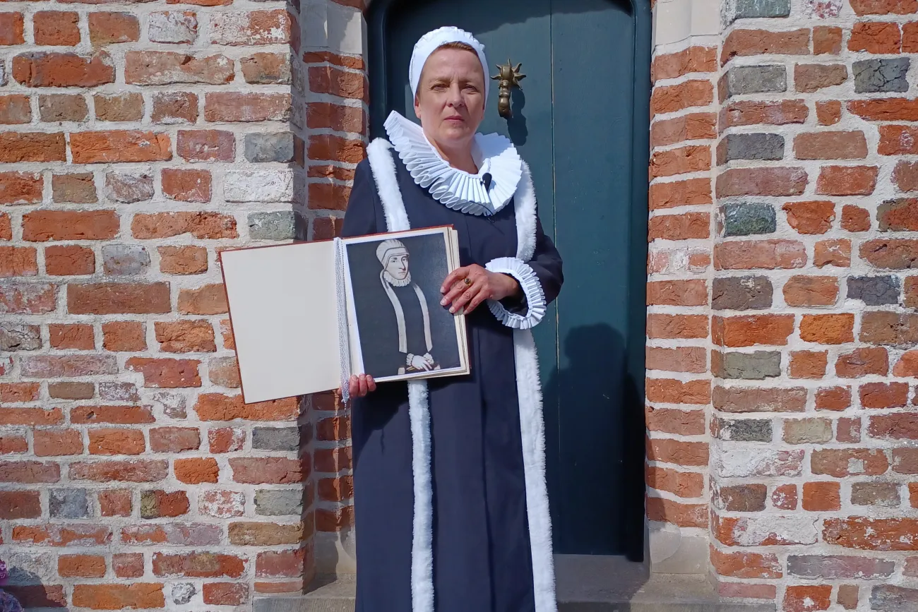 Frau in historischer Tracht hält ein Porträt von Gräfin Anna von Ostfriesland vor Backsteinmauer.