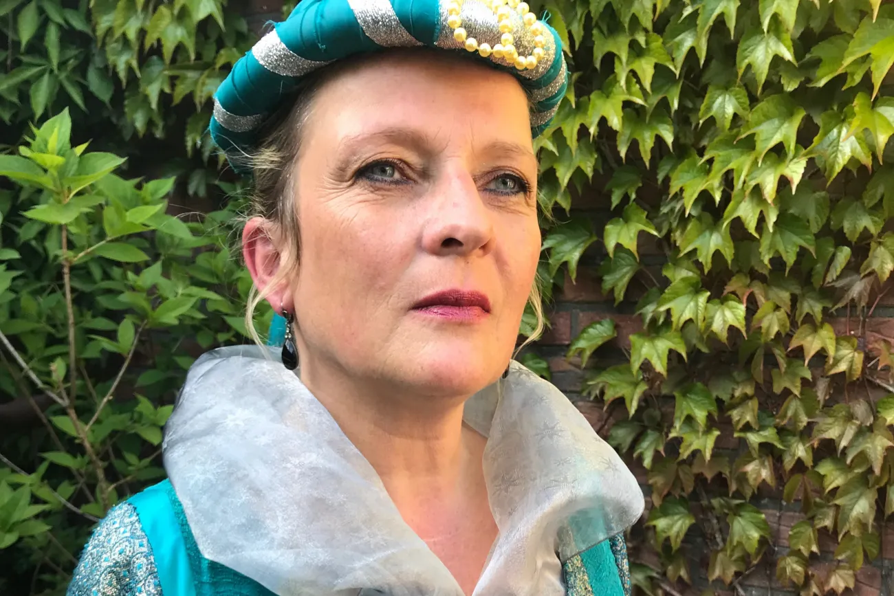 Frau in historischem Kostüm mit türkis-silberner Haube und besticktem Kleid vor grüner Efeuwand.