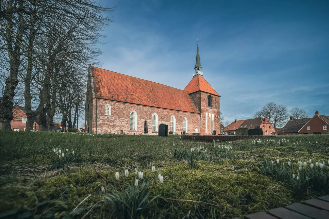 Backsteinkirche mit rotem Ziegeldach und Turm im Rundwarfendorf Rysum, umgeben von alten Bäumen.