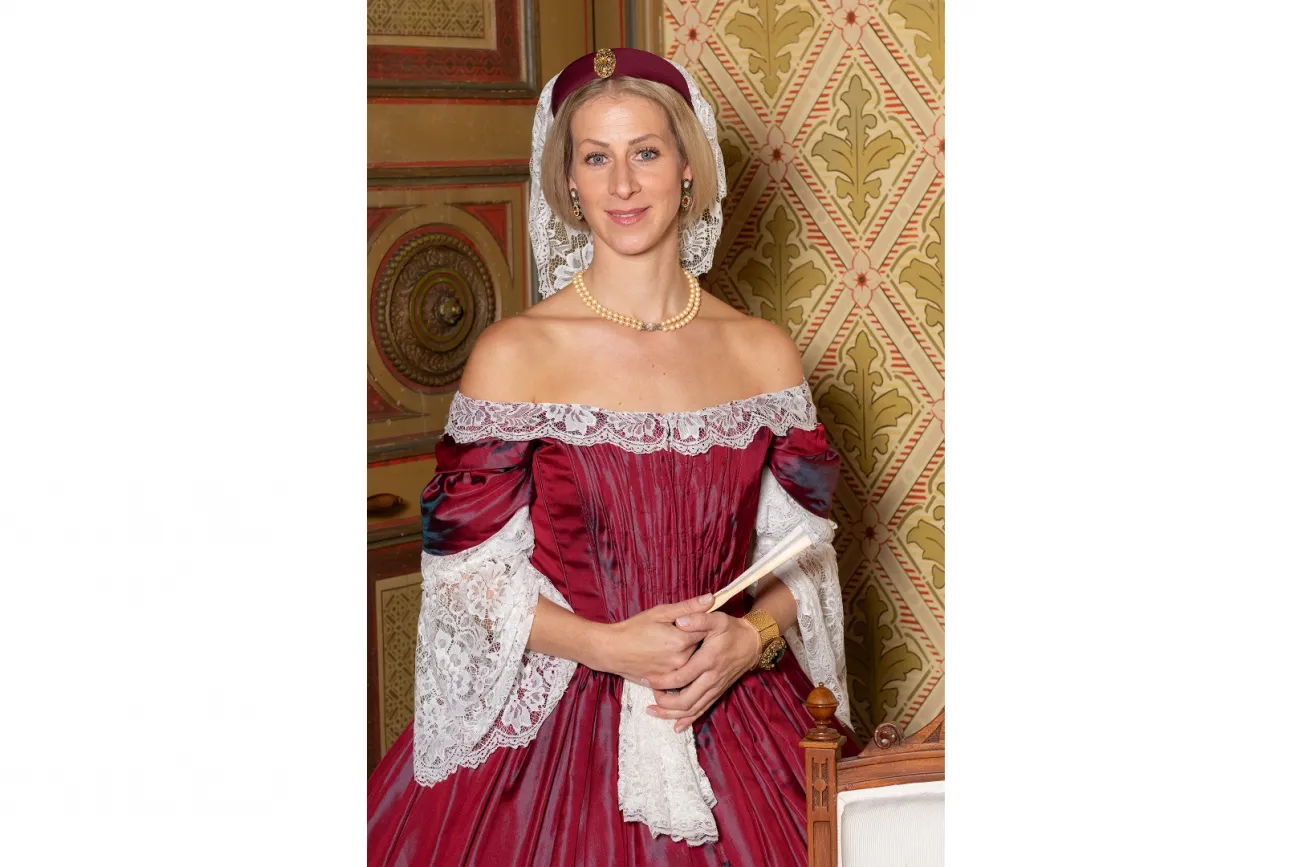 Die bestehende Beschreibung lautet: "Das Bild zeigt die Stadtführerin." Hier ist die optimierte Version: "Stadtführerin in historischer Tracht vor kunstvollem, antikem Hintergrund stehend."