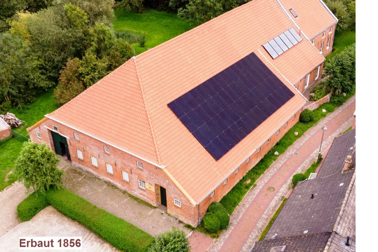 Backsteingebäude mit rotem Ziegeldach und Solaranlage, umgeben von Grünflächen und Pflasterweg.