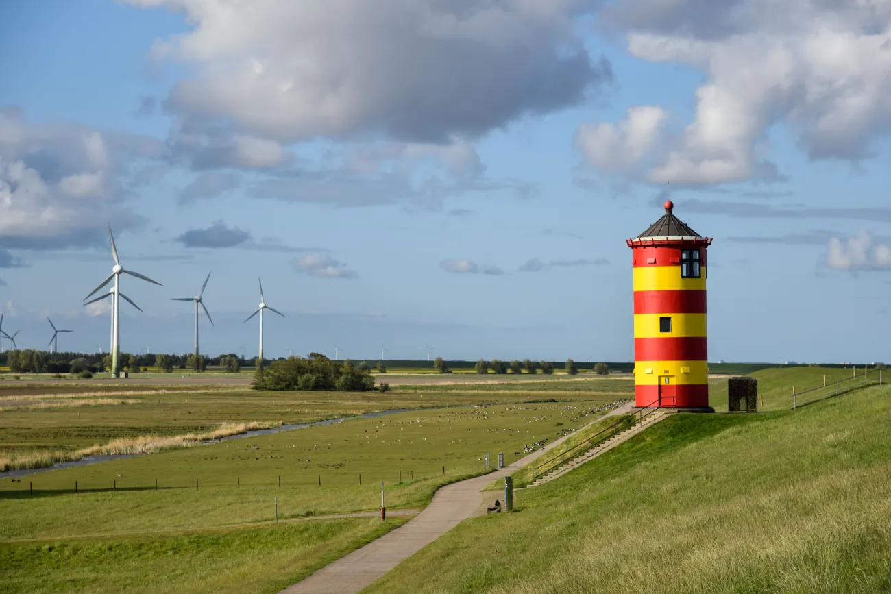 Roter und gelber Leuchtturm auf grünem Deich mit weitem Himmel und Windrädern im Hintergrund.