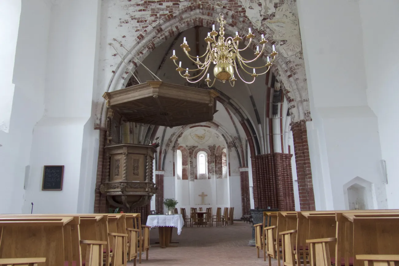 Innenansicht der Kirche Pilsum mit Holzbänken, Kanzel und Kronleuchter unter einem Backsteingewölbe.