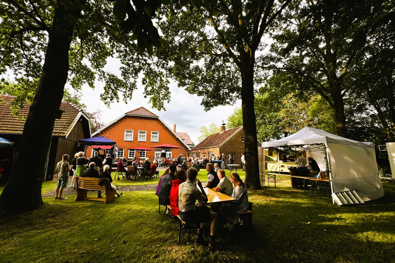 Menschen sitzen an Tischen und Bänken im Garten vor einem roten Backsteinhaus unter Bäumen bei einer Veranstaltung mit Bühne und Pavillons