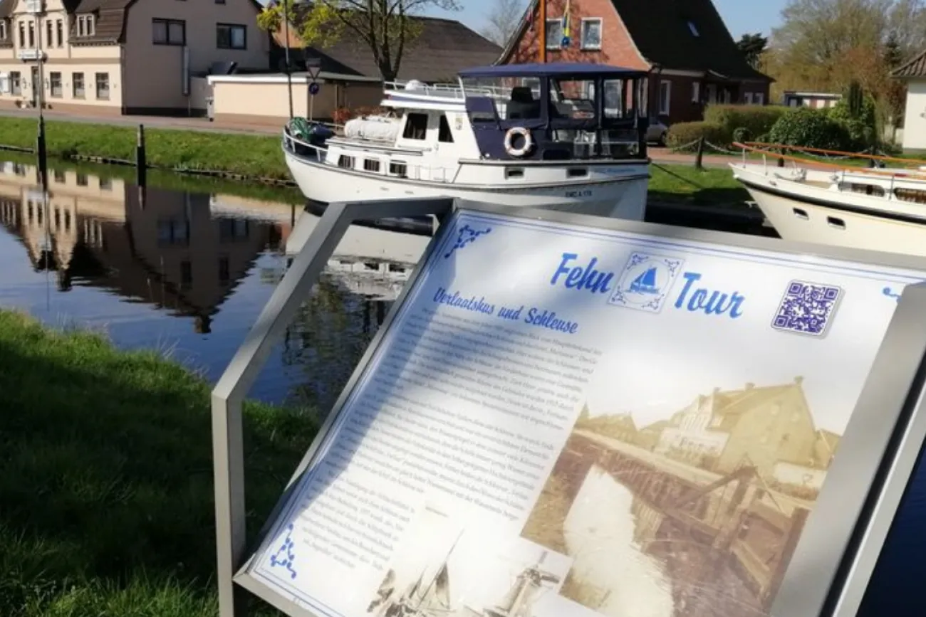 Infotafel zum historischen Rundgang vor einem Kanal mit Boot und Häusern in Rhauderfehn