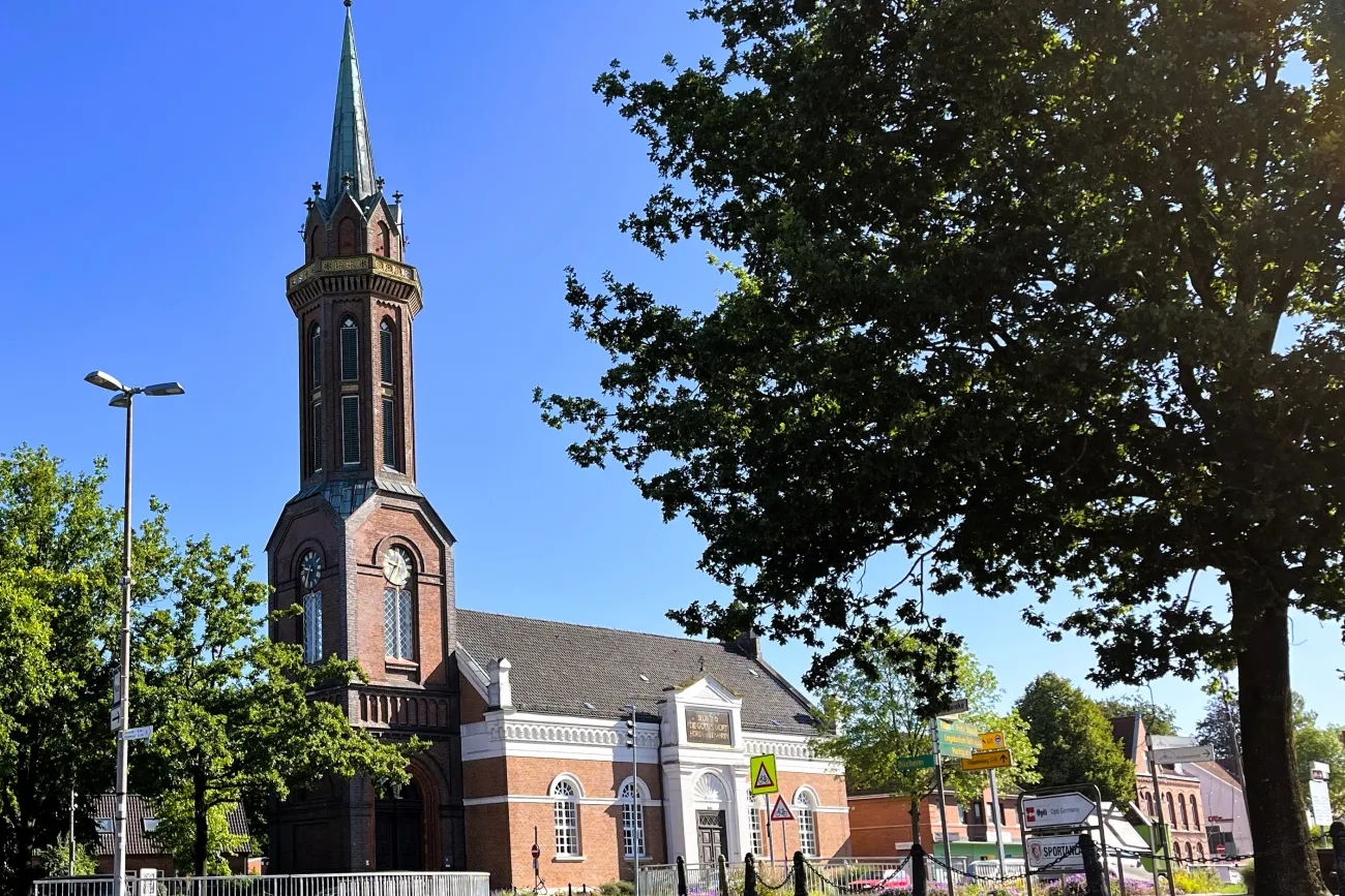 Kirchturm mit spitzem Dach neben Bäumen und einem Gebäude mit weißer Fassade unter blauem Himmel