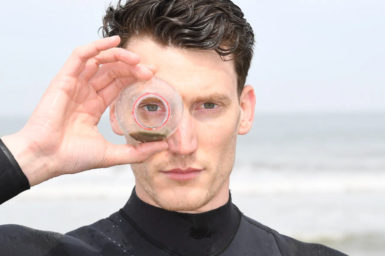 Chris Weigand im Neoprenanzug am Meer schaut durch eine Plastikflasche in die Kamera.