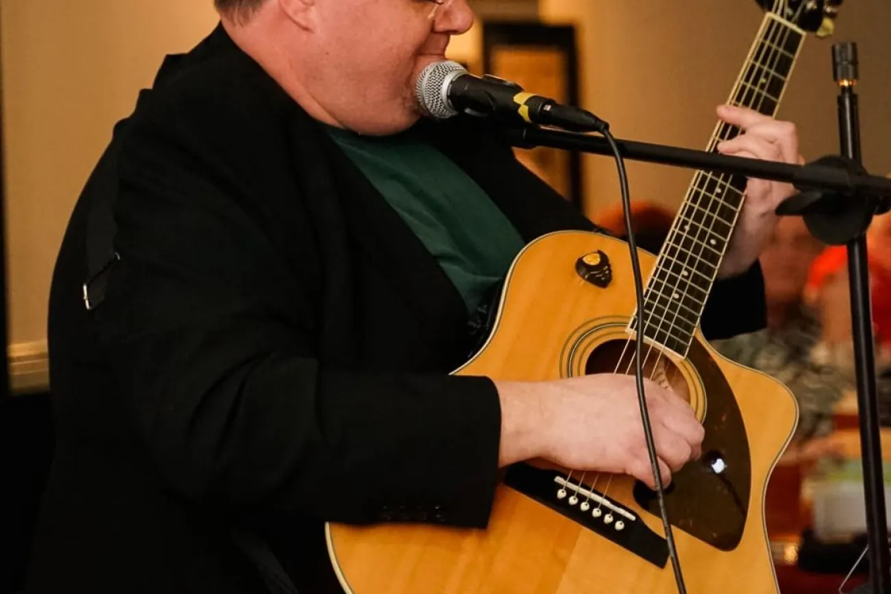Person sitzt und spielt akustische Gitarre vor Mikrofon in Innenraum mit unscharfem Hintergrund