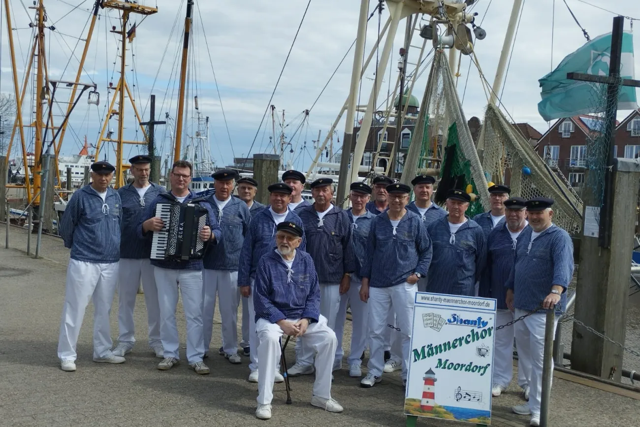 eine Gruppe von 17 Männern in blauen Seemannshemden und weißer Hose mit Seemannsmütze. Im Hintergrund sieht man Bootsmasten.