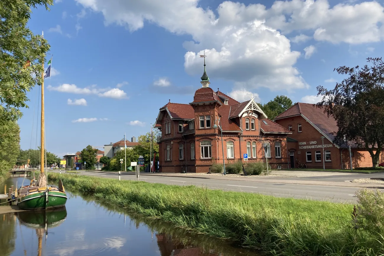 Zwei Gebäude in Backsteinbauweise, eines mit einem Turm und einem Schornstein, am Straßenrand. Im Vordergrund ein schmaler Kanal mit einem Boot.