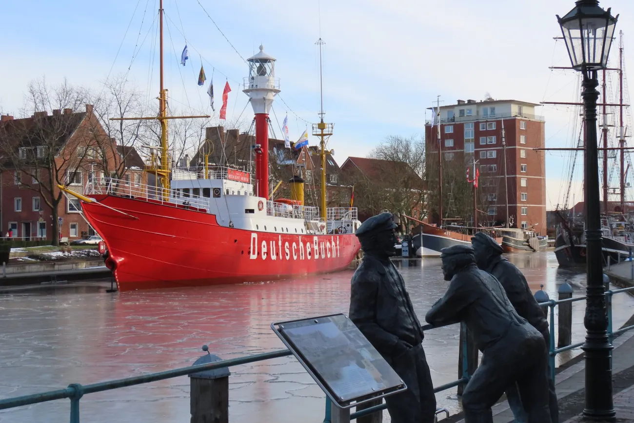 Rotes Feuerschiff mit der Aufschrift »Deutsche Bucht« im Hafen, davor zwei bronzene Figuren an einem Geländer und eine Laterne.