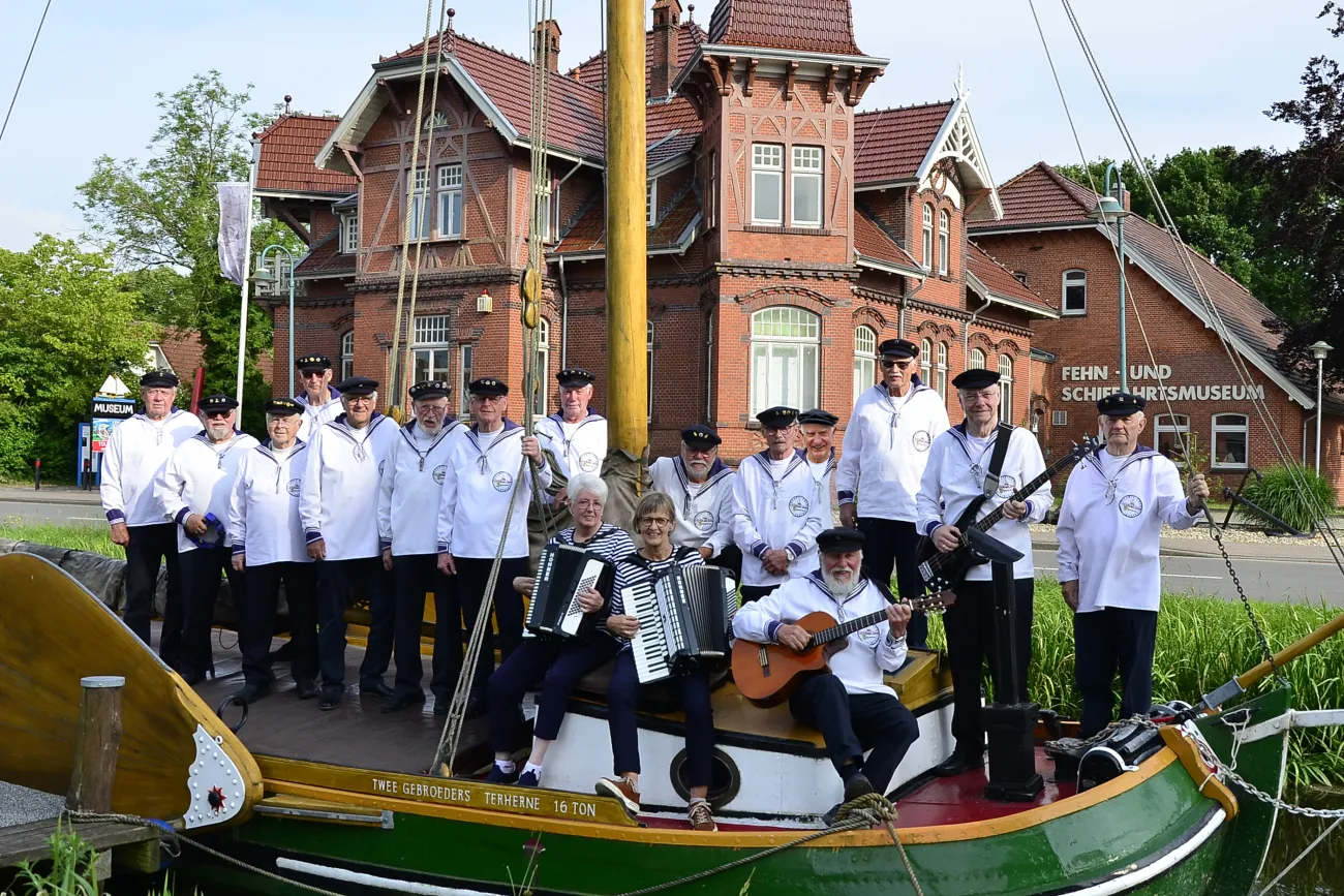 Gruppe von Männern in weißen Matrosenhemden auf einem historischen Segelboot vor einem Backsteingebäude mit der Aufschrift 'FEHN- UND SCHIFFFAHRTSMUSEUM'