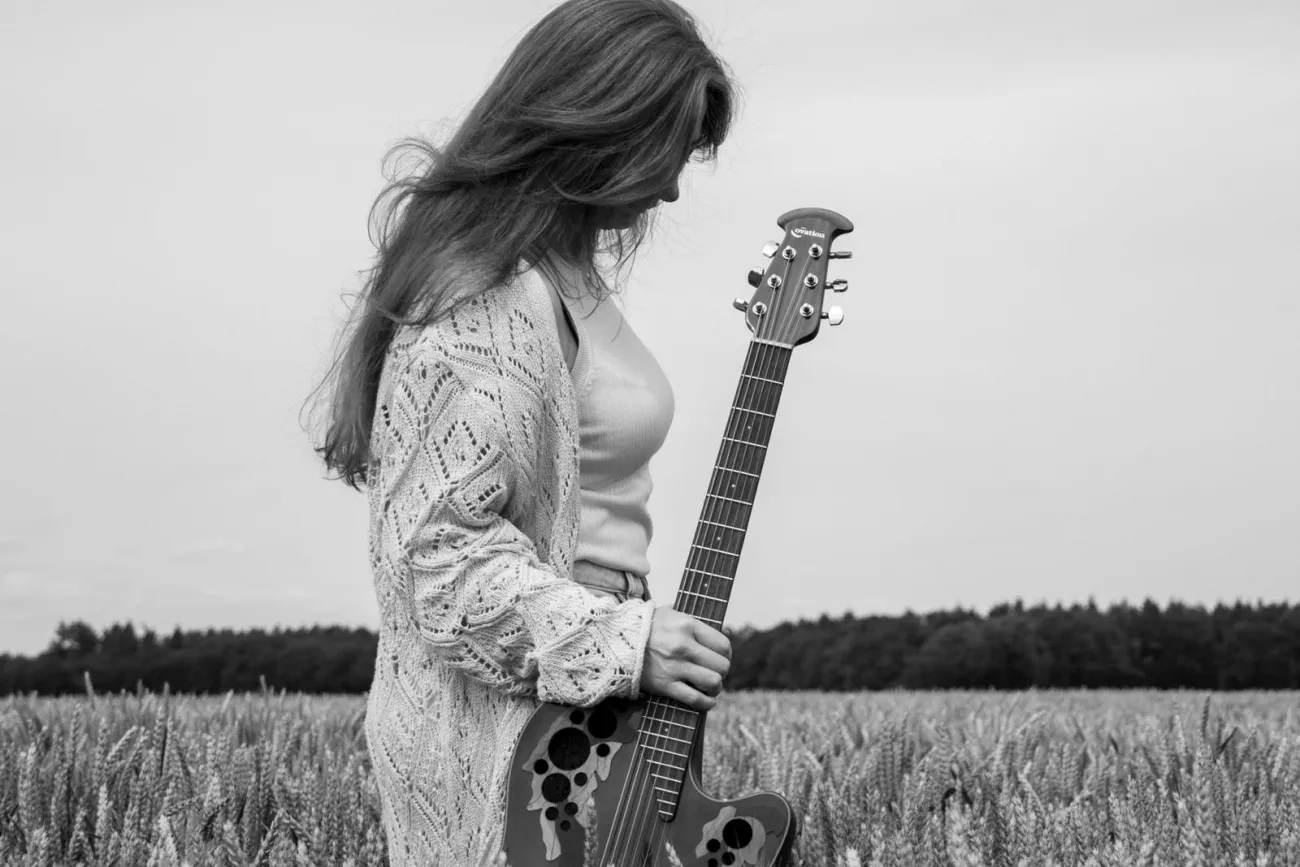 Frau mit langem Haar und Strickjacke steht in einem Getreidefeld und hält eine Akustikgitarre im Stehen