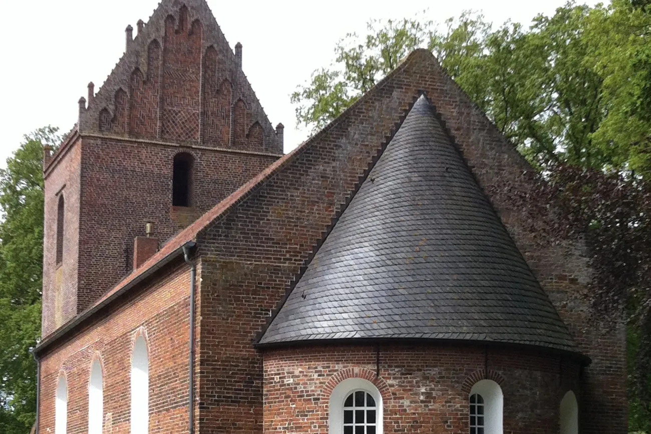 Backsteinkirche mit romanischem Rundturm und gotischem Giebel, umgeben von Bäumen