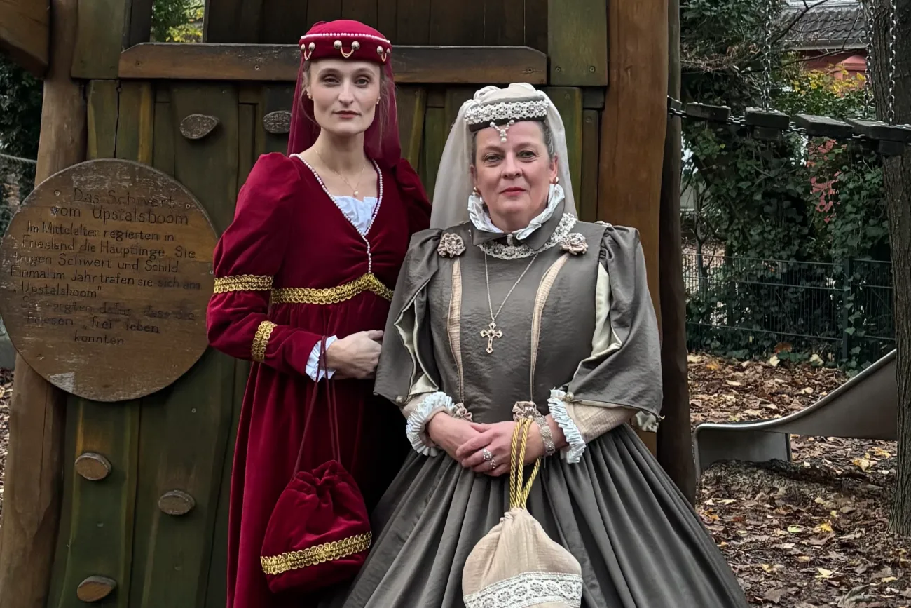 Zwei Frauen in historischen Kleidern posieren im Freien vor einer hölzernen Kulisse im Herbst.