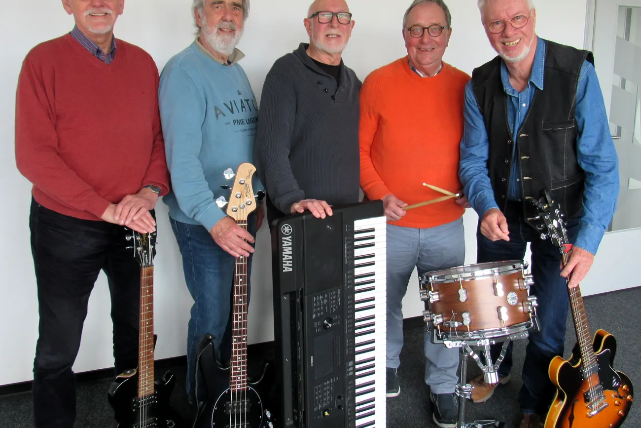 Fünf Männer stehen nebeneinander, vier halten Musikinstrumente: zwei E-Gitarren, einen E-Bass, ein Keyboard und ein Schlagzeug mit Sticks.