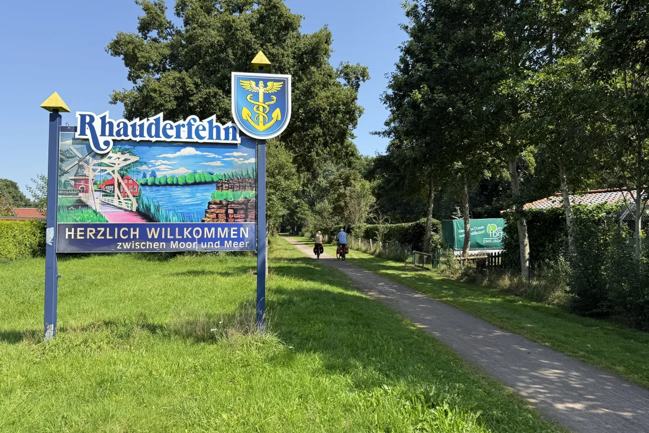 Willkommensschild mit dem Schriftzug 'Rhauderfehn' und 'Herzlich willkommen zwischen Moor und Meer' neben einem Weg mit zwei Radfahrern und Bäumen im Hintergrund
