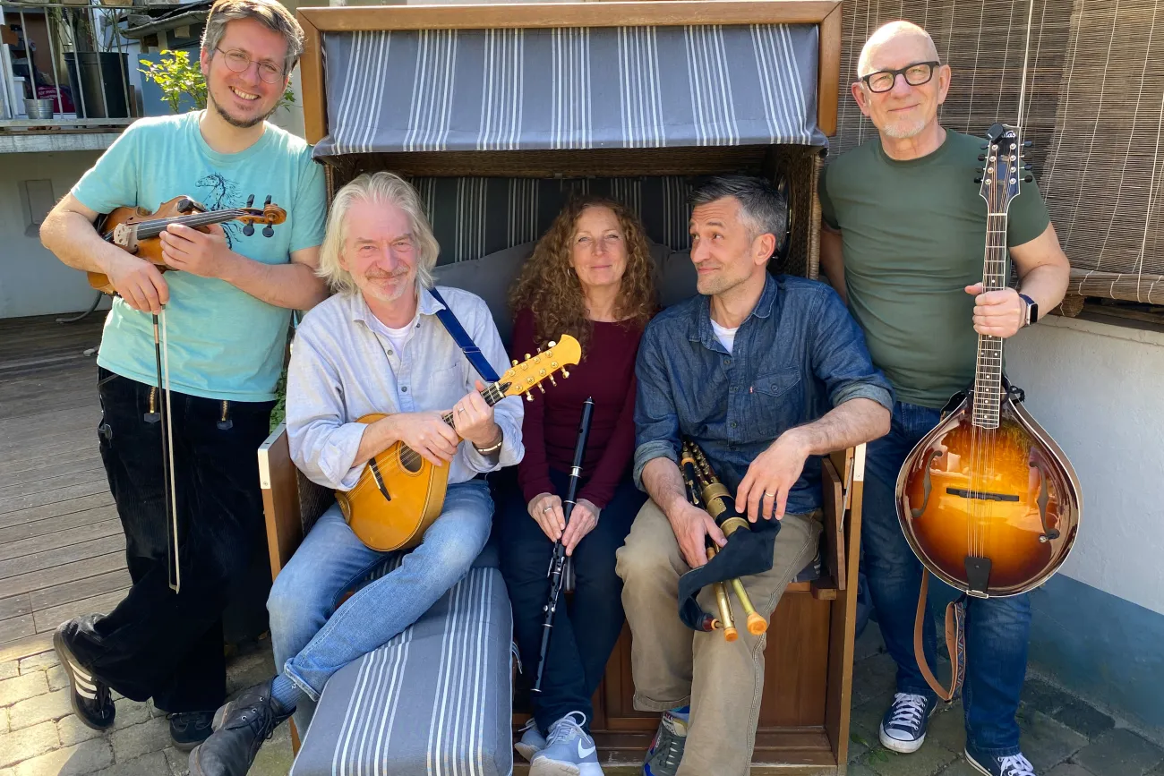Fünf Personen mit Musikinstrumenten sitzen und stehen um eine Strandkorbbank auf gepflastertem Boden.