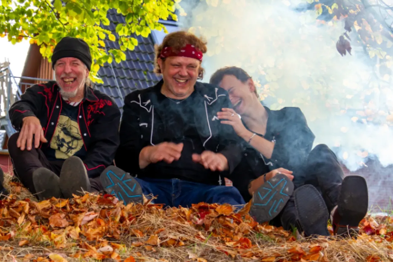 Drei Personen sitzen auf einem mit Herbstlaub bedeckten Boden vor einem Hausdach, umgeben von Rauch und Bäumen mit grünen und gelben Blättern