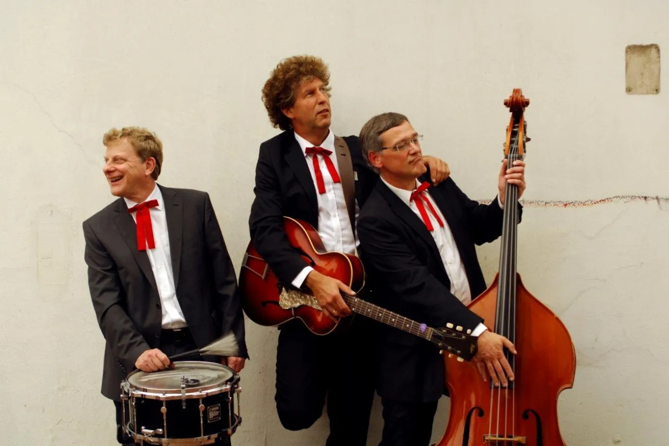 Drei Musiker im Anzug mit roter Fliege posieren mit Trommel, Gitarre und Kontrabass.