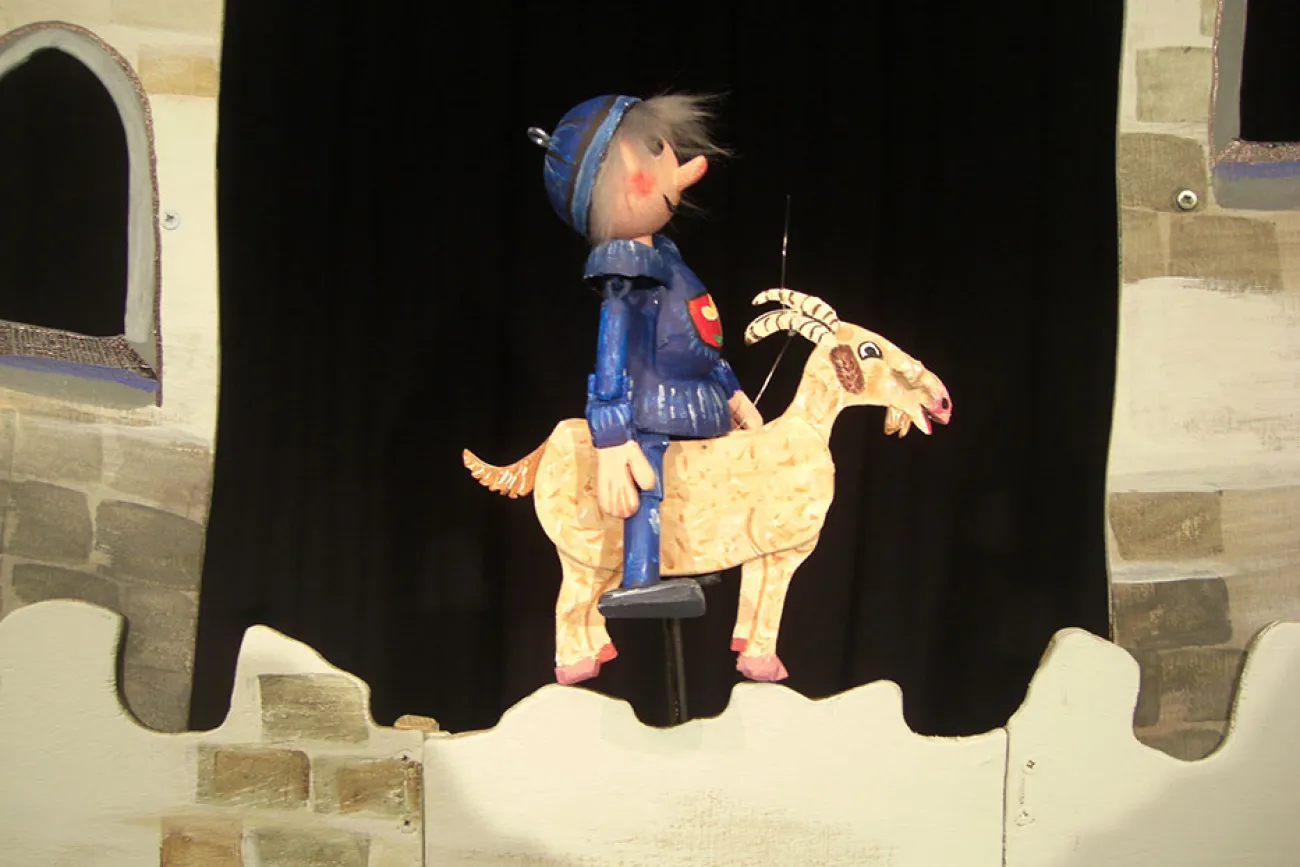 Puppentheater: Kleiner Ritter in blauer Rüstung reitet auf einer Ziege vor einer Burgmauer.