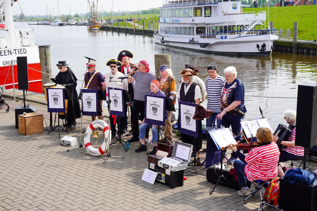 Ein Shantychor in Piratenkostümen musiziert am Hafenbecken vor einem weißen Schiff.