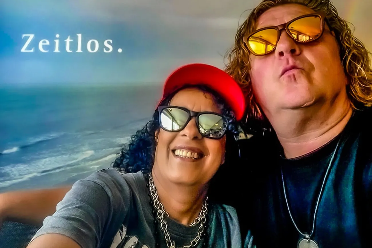 Die Musiker Gerrit und Amina als Selfie-Porträt vor einer Meereskulisse mit Schriftzug 'Zeitlos.'.