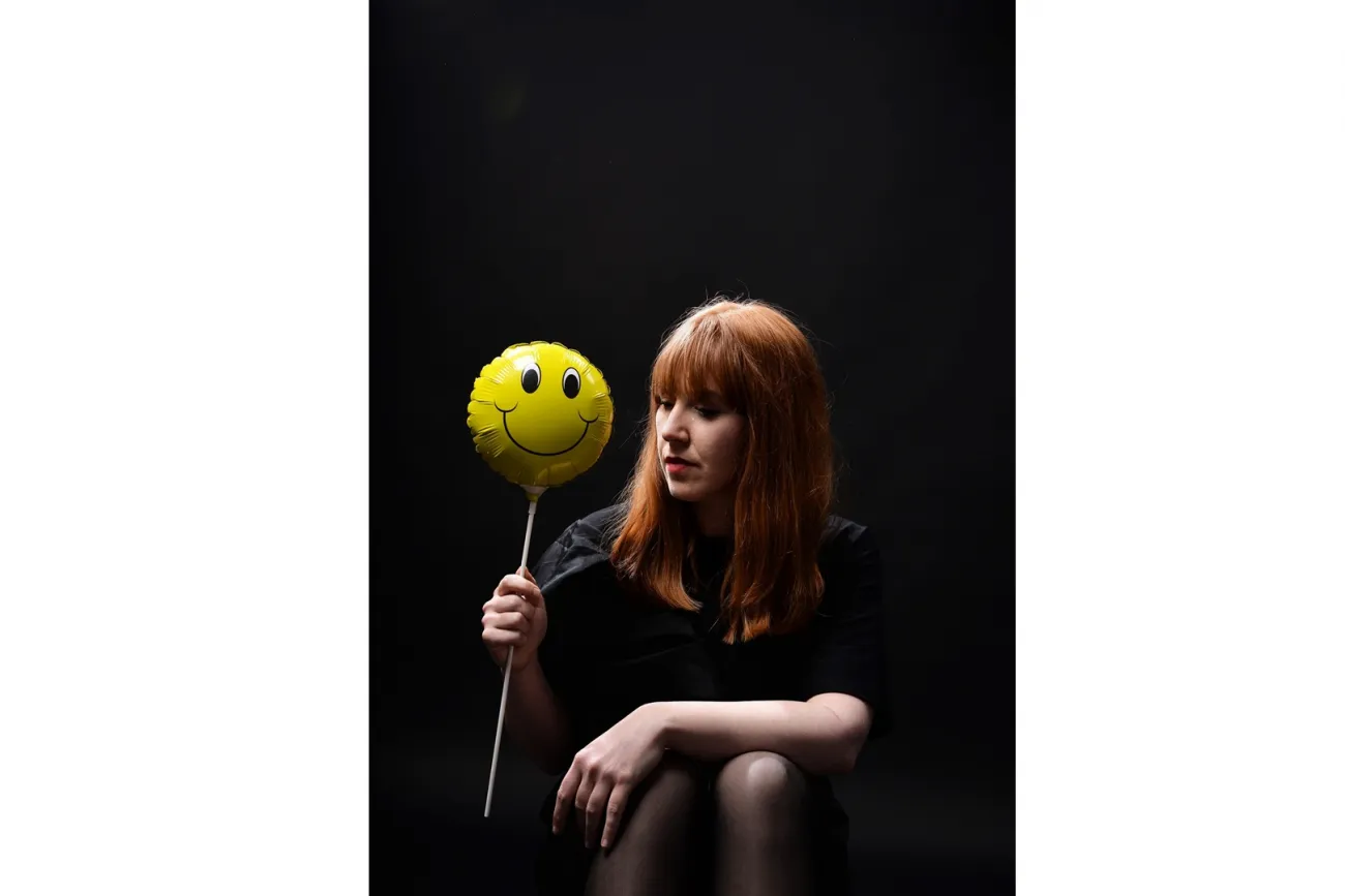 Frau mit rotem Haar hält gelben Smiley-Ballon vor dunklem Hintergrund.