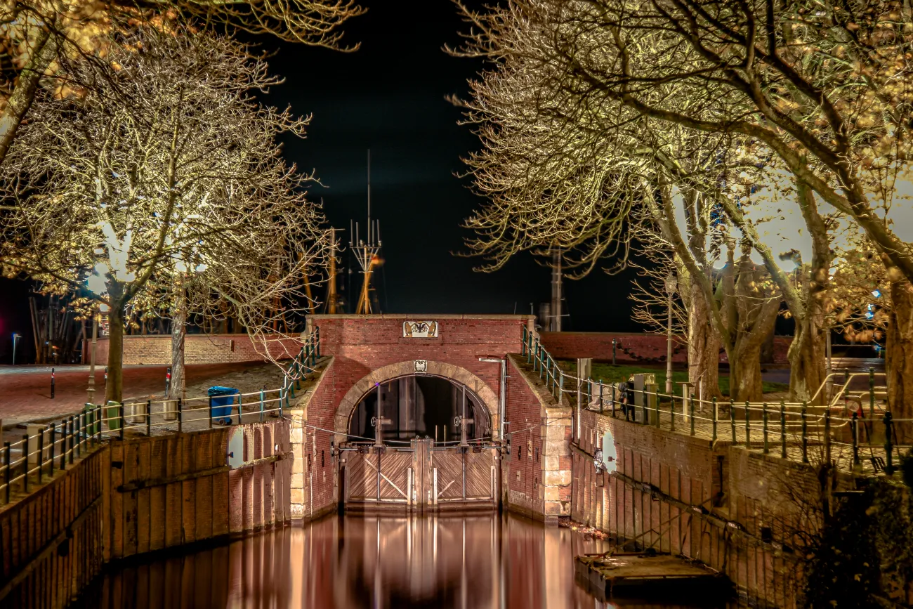 Beleuchtete Schleuse in Greetsiel, umgeben von kahlen Bäumen, spiegelt sich im ruhigen Wasser der Nacht.