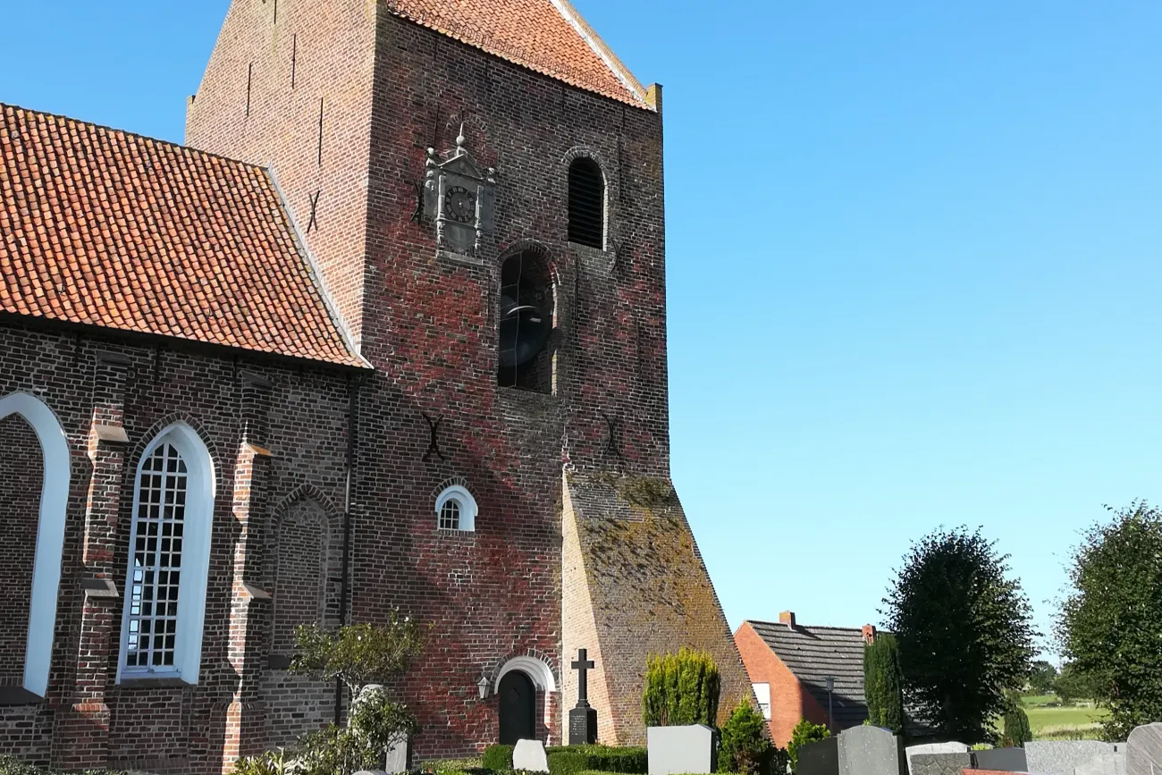 Backsteinkirche mit hohem Turm und rotem Ziegeldach vor blauem Himmel, umgeben von Grabsteinen.