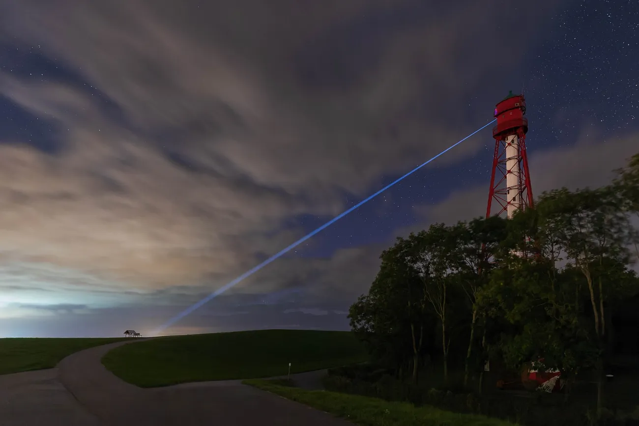 Der Leuchtturm von Campen bei Nacht. Ein dünner Lichtstrahl scheint vom Turm genau auf eine Bank gerichtet zu sein.