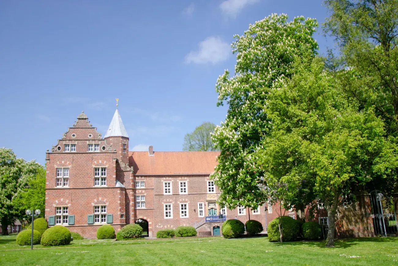 Farbfotografie der Haneburg in Leer, aufgenommen im Mai. Die historische Burganlage mit ihrer roten Ziegelfassade und den markanten Renaissance-Elementen liegt idyllisch eingebettet in einer grünen Parklandschaft. Im Vordergrund sind gepflegte Rasenflächen und blühende Sträucher zu sehen, die das Bauwerk harmonisch umrahmen. Die Haneburg ist ein bekanntes Kultur- und Bildungszentrum der Stadt Leer und beherbergt heute die Volkshochschule, die dort Veranstaltungen und Kurse anbietet