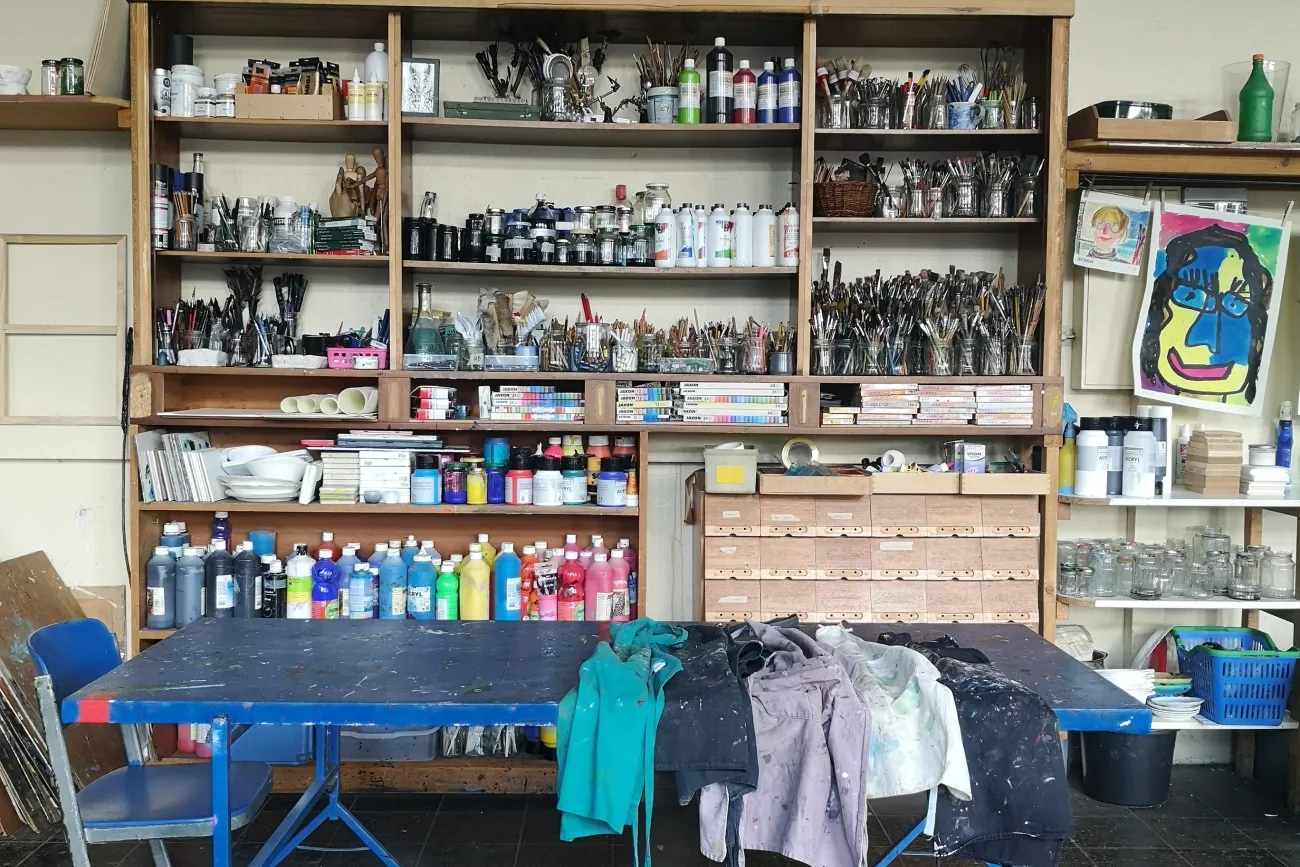 Regal in der Kunstschule mit Pinseln und Farben, davor ein Tisch mit Stühlen