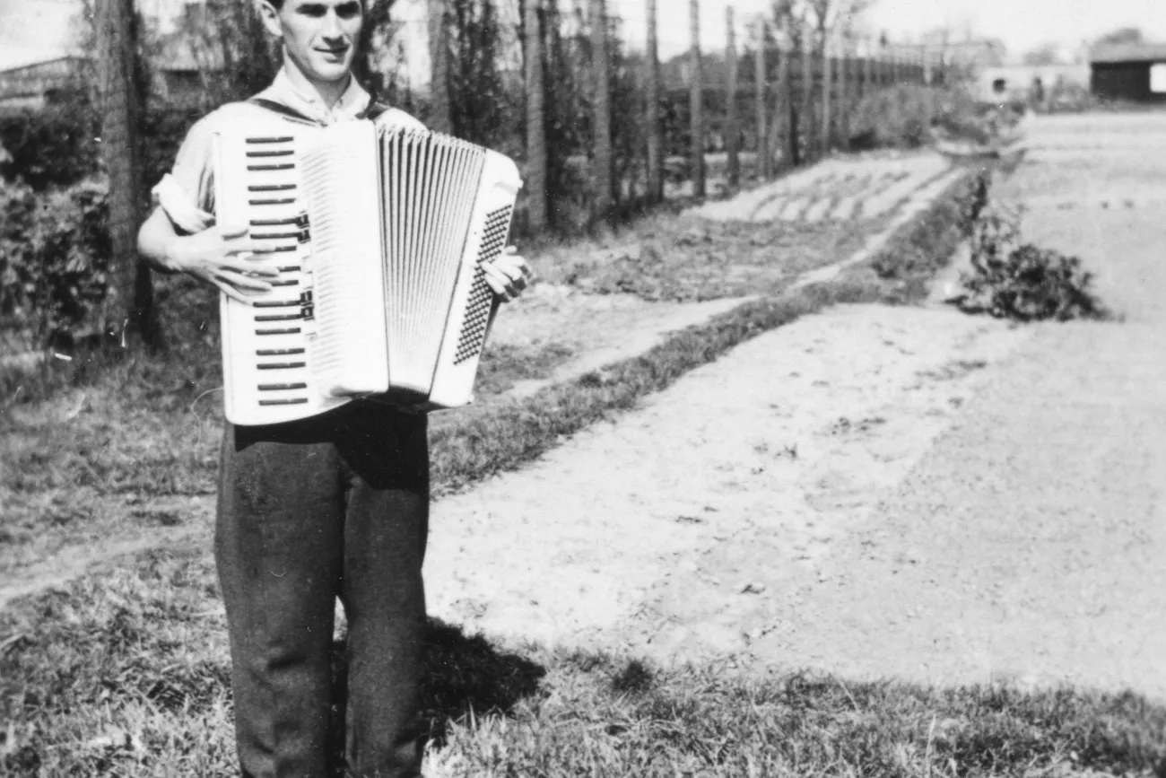 Ein Mann spielt Akkordeon vor einem Stacheldrahtzaun. Das Bild ist in Schwarzweiß. 