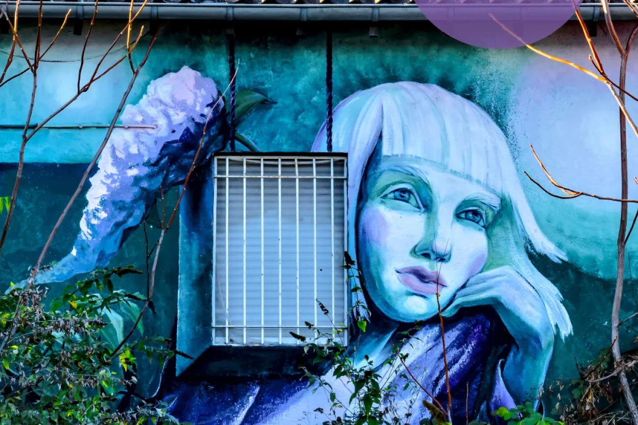 Eine Einladung zur Fotoausstellung des Fotoforums. Das Bild zeigt ein farbenfrohes Wandgemälde mit einem surrealen Porträt einer Frau mit weißem Haar, die nachdenklich auf ihre Hand gestützt ist. Das Fenster mit Gitter integriert sich in das Kunstwerk. Pflanzen wachsen davor, teils mit gelblichen Blättern.