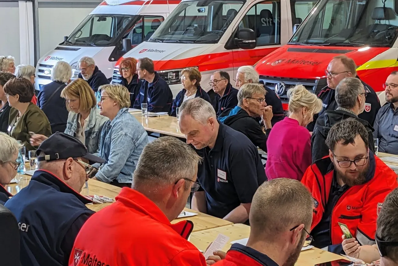 Eine Gruppe von Maltesern sitzt anlässlich eines Ehrenamtstages vor Einsatzfahrzeugen auf Bierzeltgarnituren