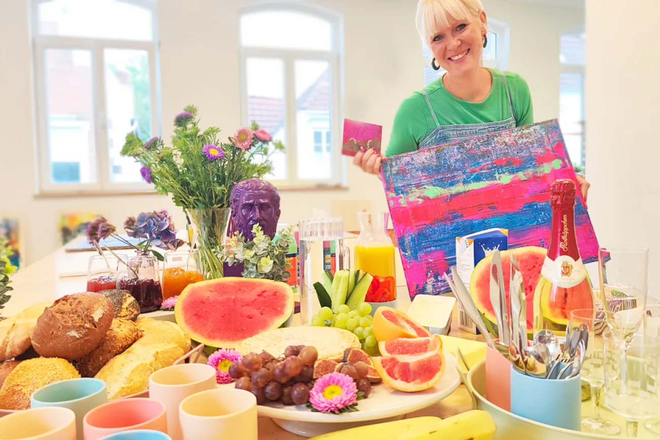 Frau mit grünem Pullover und einem selbstmaltem Bild in der Hand vor einem gedeckten Frühstückstisch
