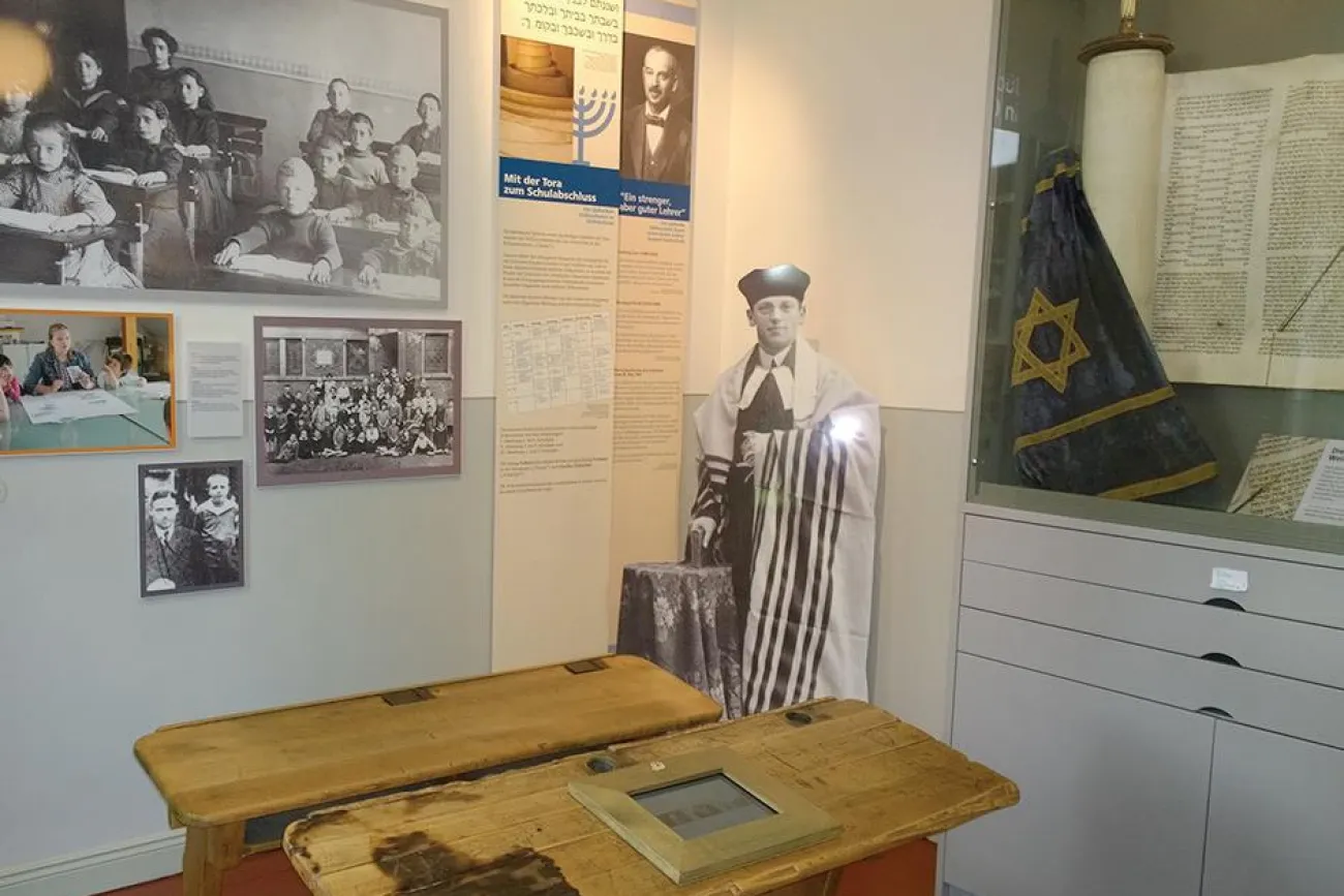 Im Vordergrund stehen zwei historische Schulbänke. Im Hintergrund ist eine Torarolle zu sehen sowie Fotos aus historischen Klassenzimmern und ein Fotoaufsteller des vorletzten Lehrers der Gemeinde August-Gottschalk