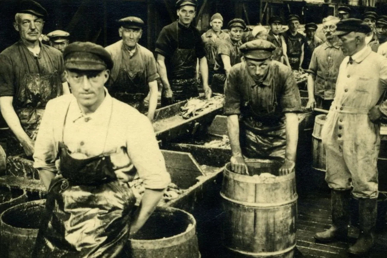 Man sieht Männer in Arbeitskleidung vermutlich an Deck eines Loggers. Die männer schauen in die Kamera und halten mit der Arbeit des Kehlens und Verpackens inne.