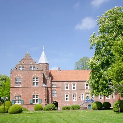 Farbfotografie der Haneburg in Leer, aufgenommen im Mai. Die historische Burganlage mit ihrer roten Ziegelfassade und den markanten Renaissance-Elementen liegt idyllisch eingebettet in einer grünen Parklandschaft. Im Vordergrund sind gepflegte Rasenflächen und blühende Sträucher zu sehen, die das Bauwerk harmonisch umrahmen. Die Haneburg ist ein bekanntes Kultur- und Bildungszentrum der Stadt Leer und beherbergt heute die Volkshochschule, die dort Veranstaltungen und Kurse anbietet