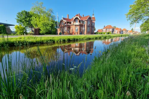 Rotes Backsteinmuseum spiegelt sich im Wasser des Rajenkanals hinter grünem Ufergras.