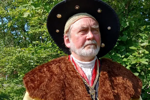 Mann in historischer Kleidung mit Pelzkragen und großem Hut vor grünem Hintergrund im Freien.