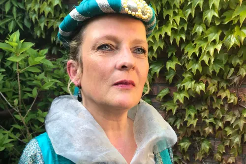 Frau in historischem Kostüm mit türkis-silberner Haube und besticktem Kleid vor grüner Efeuwand.