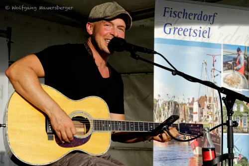 Musiker spielt lächelnd Gitarre und singt bei einem Sommerkonzert in Greetsiel auf einer Open-Air-Bühne.