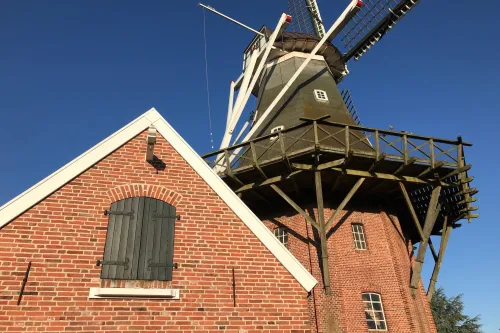 Historische Windmühle aus rotem Backstein mit Holzgalerie und weißen Flügeln unter blauem Himmel.