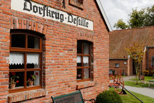 altes Backsteinhaus mit 2 Fenstern, an der Hauswand ist ein altes Schild "Dorfkrug Destille" angebracht, davor steht eine grüne Holzsitzbank