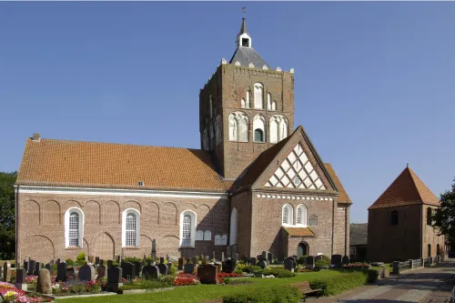 Imposante Kreuzkirche Pilsum mit massivem Vierungsturm und Friedhof unter blauem Himmel.