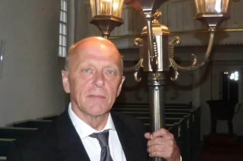 Ein Mann in Anzug hält eine historische Straßenlaterne in einem Kirchenraum mit warmer Beleuchtung.